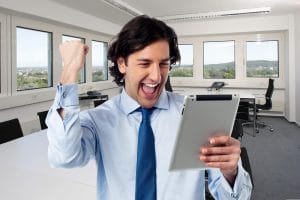 Man in a blue shirt and tie raises a fist while looking at a tablet in a bright office setting&nbsp;Hispamer Noticias