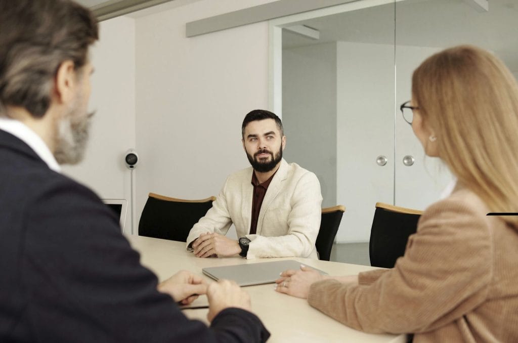 Man in a light blazer sits at a conference table facing two colleagues during a meeting in a bright office.&nbsp;Hispamer Noticias