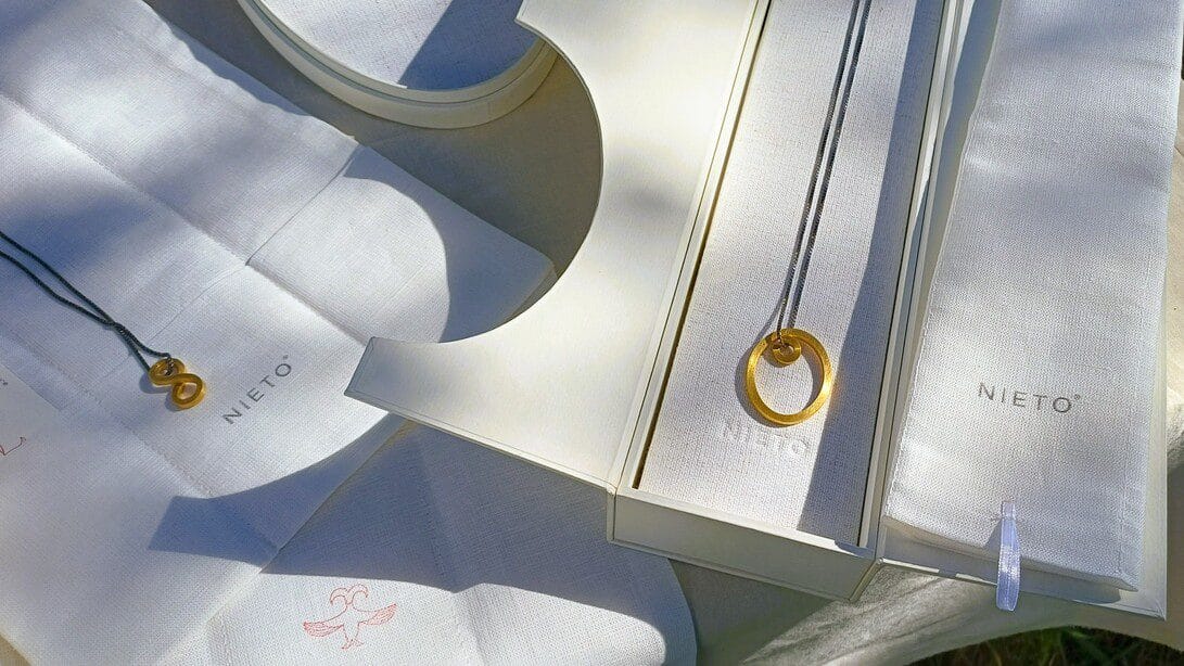 Gold circular pendant on a thin chain and two small intertwined-loop pendants displayed on white Nieto jewelry boxes outdoors.&nbsp;Hispamer Noticias