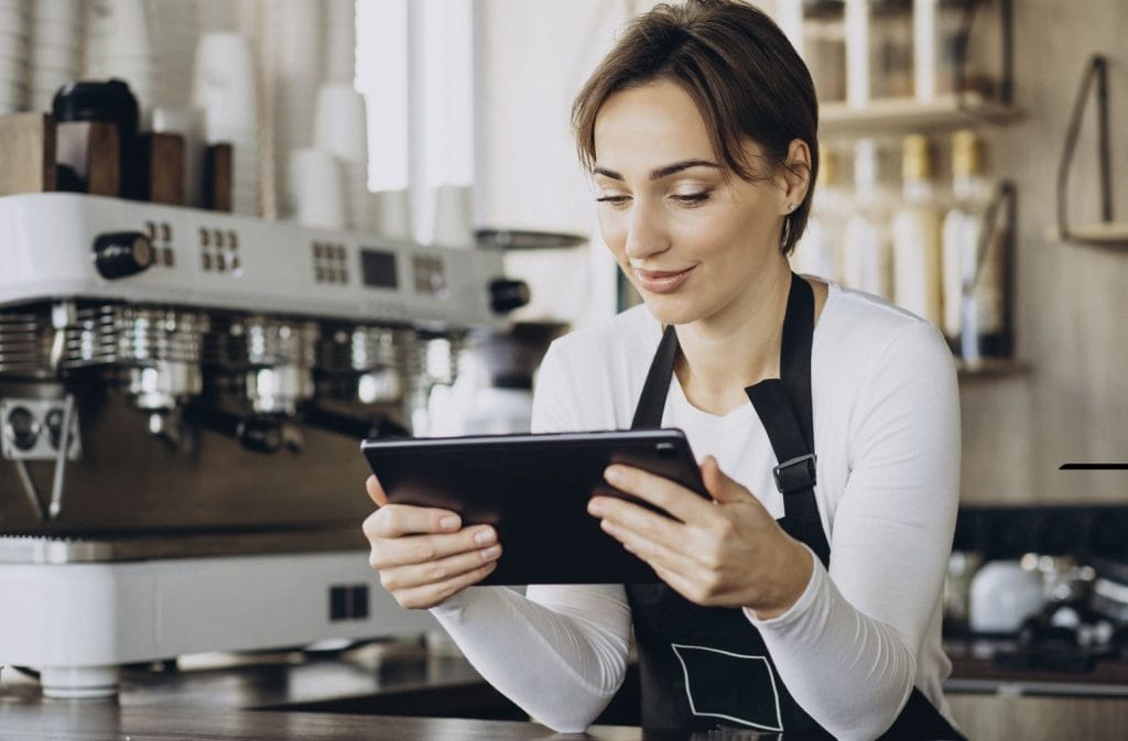 Barista in a white shirt and black apron uses a tablet at a cafe counter beside a large espresso machine.&nbsp;Hispamer Noticias