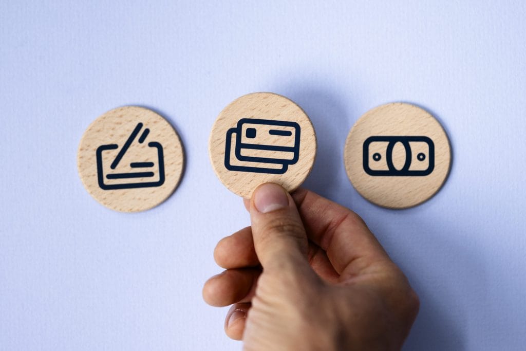 Three wooden tokens with payment icons (card left, stacked cards center, wallet right); a hand holds the center token.
