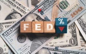 Wooden blocks spell FED with a blue percent tile and a red downward arrow, set against a backdrop of U.S. dollar bills—illustrating Federal Reserve rate changes.&nbsp;Hispamer Noticias