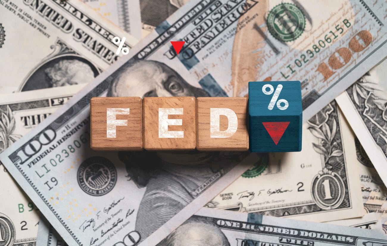 Wooden blocks spell FED with a blue percent tile and a red downward arrow, set against a backdrop of U.S. dollar bills—illustrating Federal Reserve rate changes.&nbsp;Hispamer Noticias
