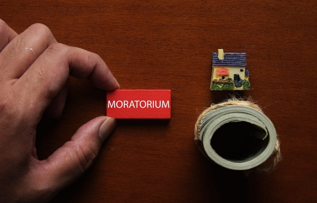 Close-up of a hand placing a red MORATORIUM block on a wooden desk, with a tiny house model and a roll of tape nearby.&nbsp;Hispamer Noticias