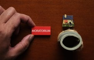 Close-up of a hand placing a red MORATORIUM block on a wooden desk, with a tiny house model and a roll of tape nearby.&nbsp;Hispamer Noticias