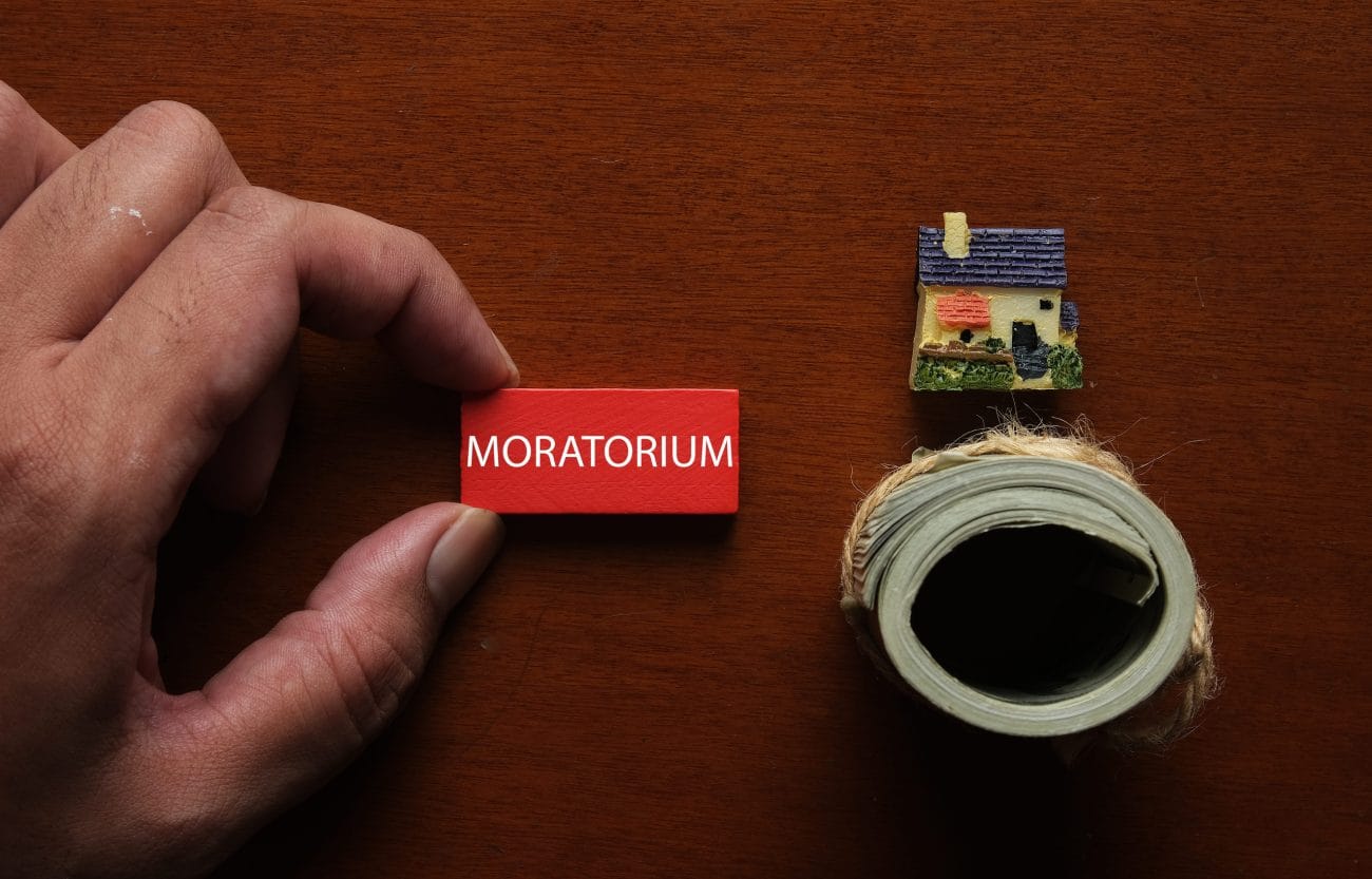 Close-up of a hand placing a red MORATORIUM block on a wooden desk, with a tiny house model and a roll of tape nearby.&nbsp;Hispamer Noticias