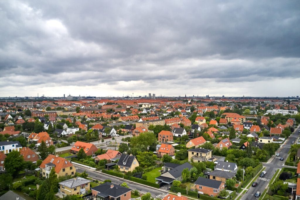 Aerial view of a dense suburban neighborhood with red-tiled roofs, green yards, and tree-lined streets under a cloudy sky in the distance.&nbsp;Hispamer Noticias
