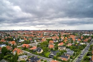 Aerial view of a dense suburban neighborhood with red-tiled roofs, green yards, and tree-lined streets under a cloudy sky in the distance.&nbsp;Hispamer Noticias