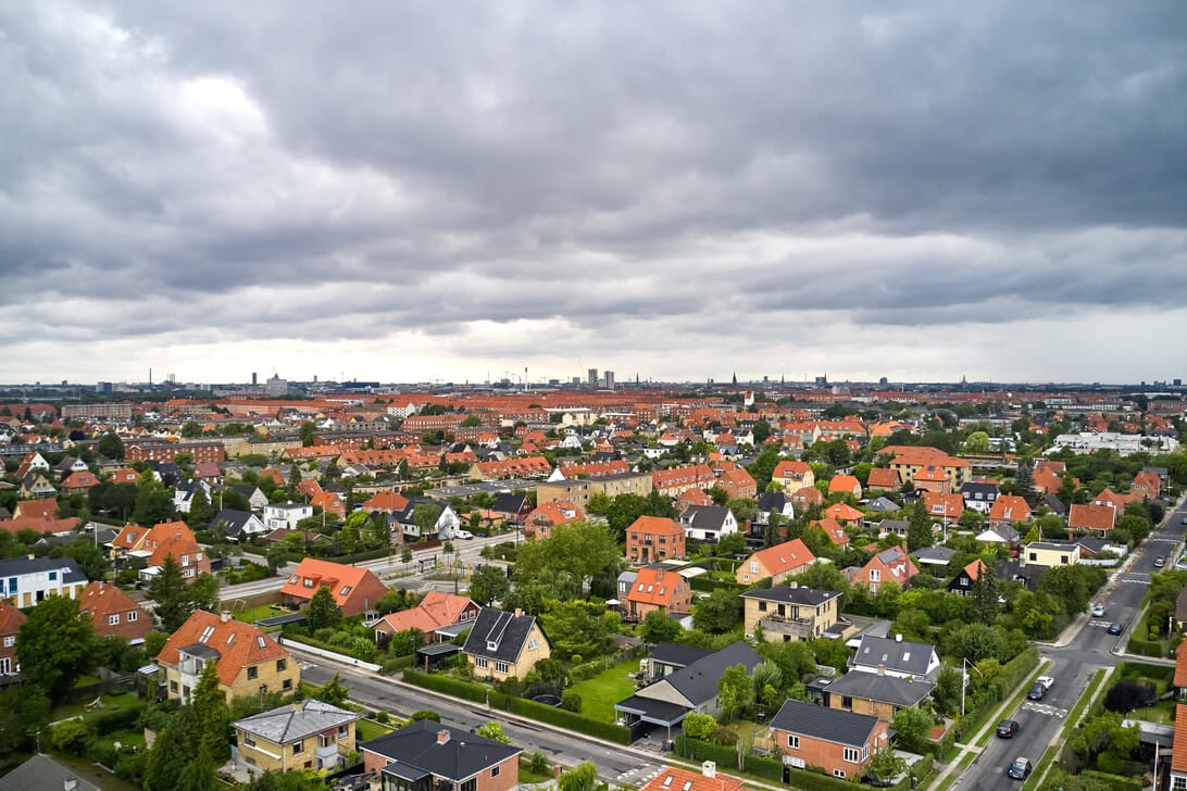 Aerial view of a dense suburban neighborhood with red-tiled roofs, green yards, and tree-lined streets under a cloudy sky in the distance.&nbsp;Hispamer Noticias