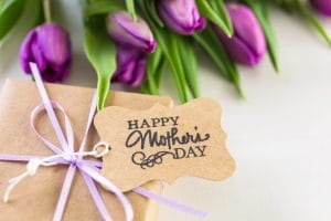 Gift-wrapped box with lavender ribbon and a kraft tag that says 'Happy Mother's Day', with purple tulips in the background.&nbsp;Hispamer Noticias