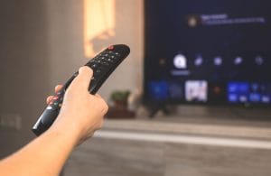 Hand holding a black TV remote aimed at a blurred wall-mounted screen in the background.&nbsp;Hispamer Noticias