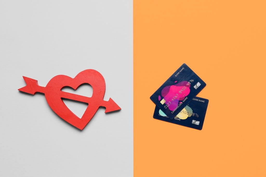 Split image: left side shows a red heart with arrows through it on a light gray background, right side shows stacked blue credit cards on an orange background, suggesting love versus money.&nbsp;Hispamer Noticias