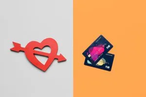 Split image: left side shows a red heart with arrows through it on a light gray background, right side shows stacked blue credit cards on an orange background, suggesting love versus money.&nbsp;Hispamer Noticias