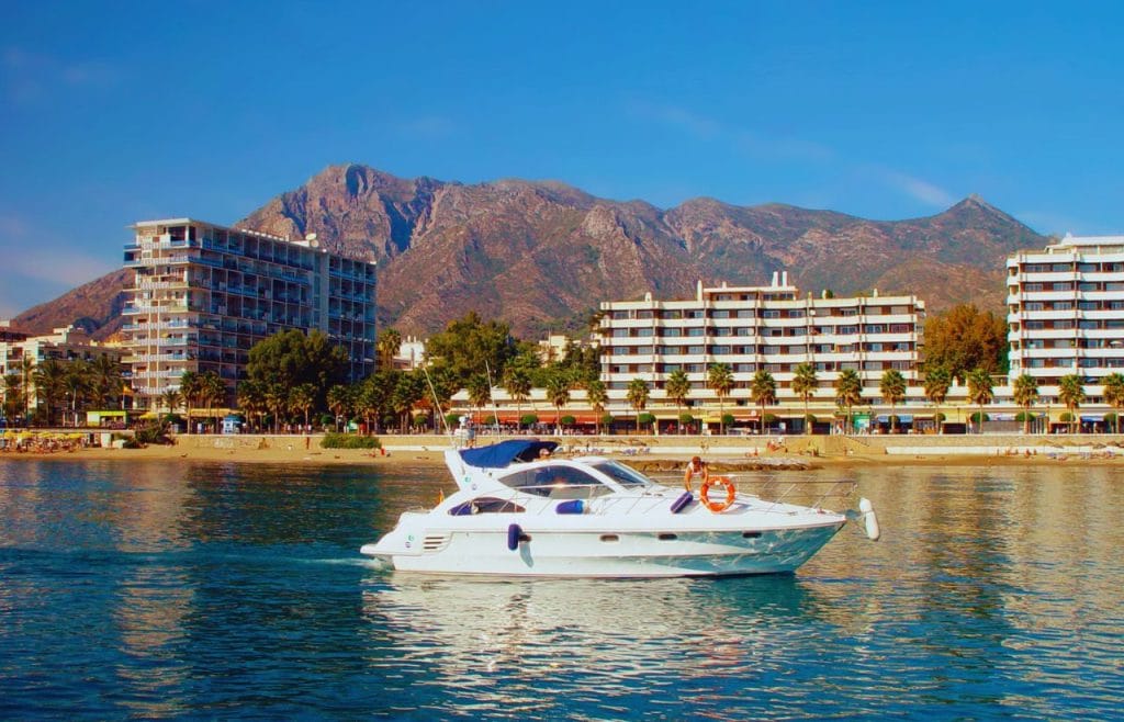 White motor yacht cruising on calm blue water with a beach and palm-lined hotels behind it, and mountains in the distance.&nbsp;Hispamer Noticias
