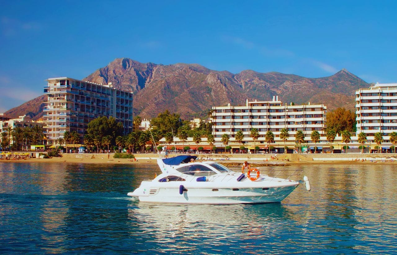 White motor yacht cruising on calm blue water with a beach and palm-lined hotels behind it, and mountains in the distance.&nbsp;Hispamer Noticias