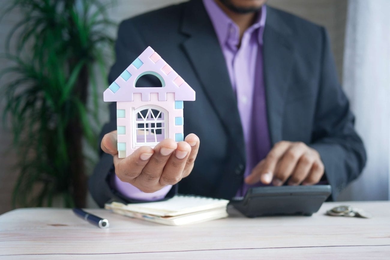 Businessperson in a suit presenting a pastel toy house over a desk with a calculator.&nbsp;Hispamer Noticias