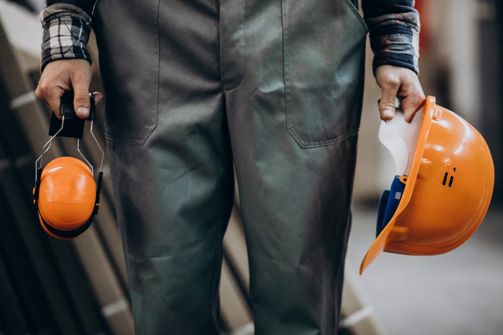 Construction worker holding orange hard hat in one hand and orange earmuffs in the other, wearing gray work overalls in a workshop setting.&nbsp;Hispamer Noticias