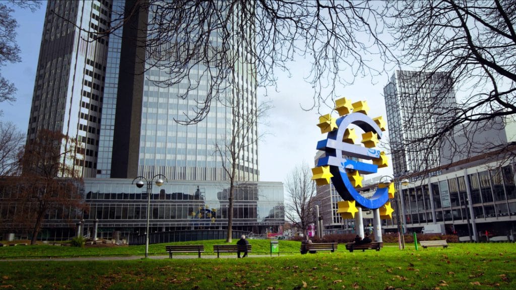 A large blue euro sign sculpture with yellow stars in a city park, with tall modern buildings in the background and a few benches nearby.&nbsp;Hispamer Noticias
