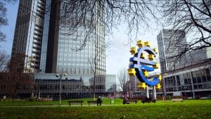 A large blue euro sign sculpture with yellow stars in a city park, with tall modern buildings in the background and a few benches nearby.&nbsp;Hispamer Noticias