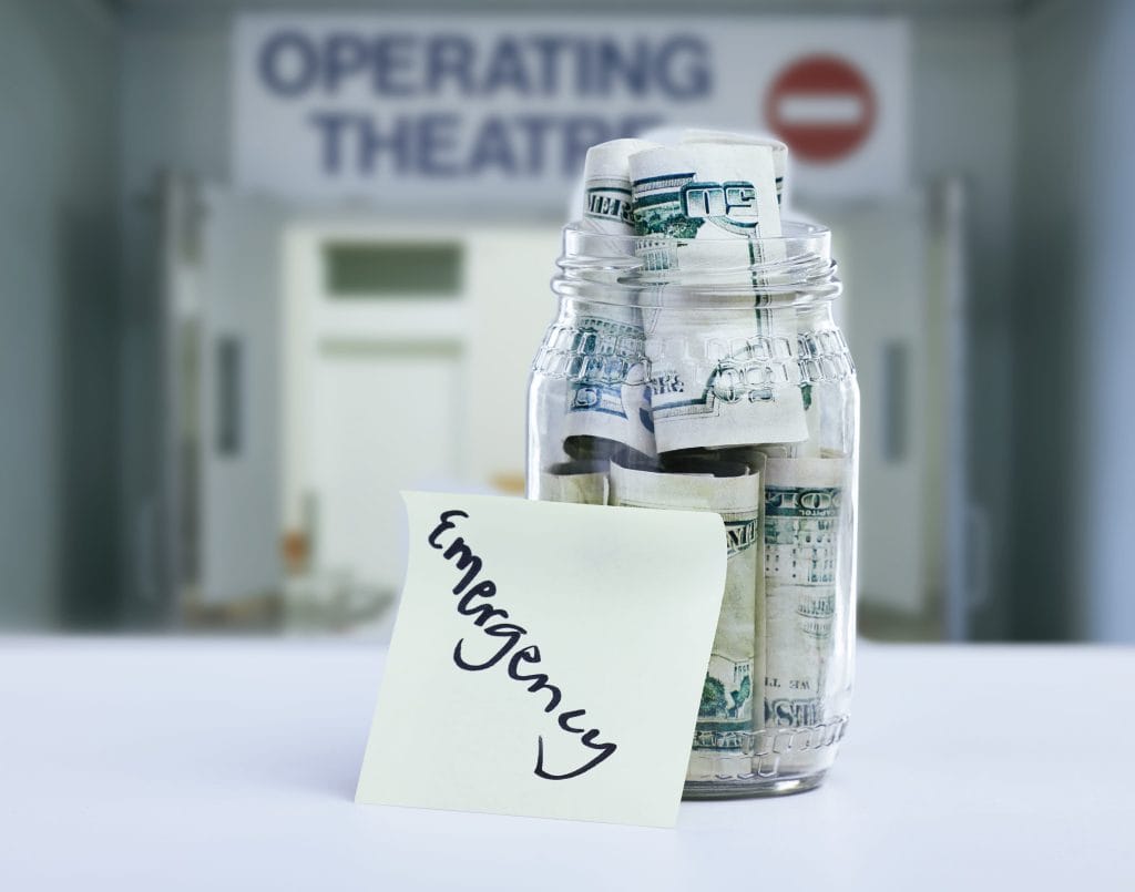 Jar filled with rolled dollar bills with an 'Emergency' note in front; a blurred hospital/theatre sign is in the background.&nbsp;Hispamer Noticias