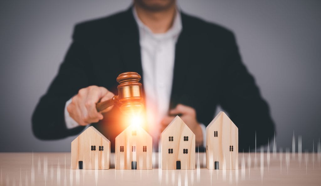 Businessperson in a suit uses a gavel to auction miniature wooden houses on a desk.&nbsp;Hispamer Noticias