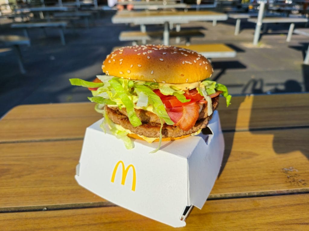 Burger with sesame bun, lettuce, tomato, cheese, and bacon on a white McDonald's box outdoors on a wooden table.&nbsp;Hispamer Noticias