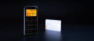 Smartphone screen showing a yellow credit card in a finance app with a transaction list; a blurred card sits in the background on a dark surface.&nbsp;Hispamer Noticias