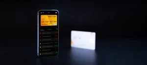 Smartphone screen showing a yellow credit card in a finance app with a transaction list; a blurred card sits in the background on a dark surface.&nbsp;Hispamer Noticias