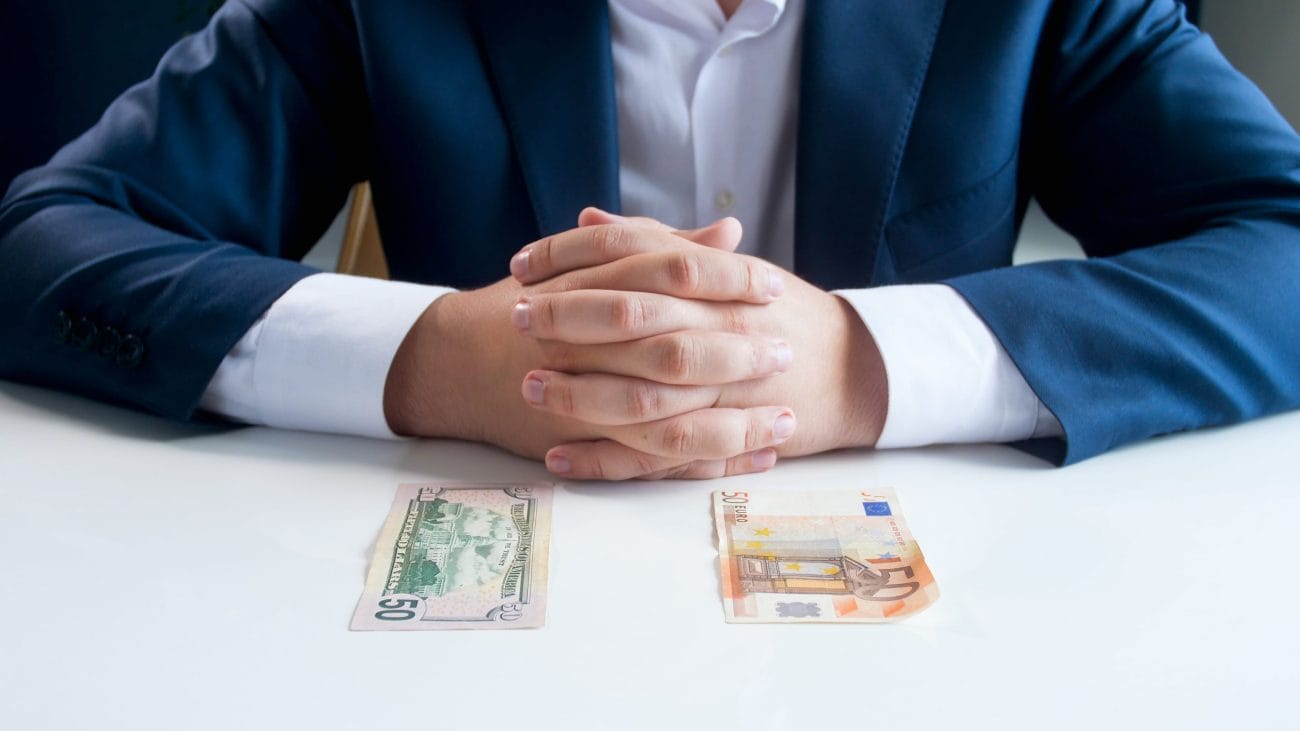 Businessperson in a blue suit with hands clasped on a table, two currency notes ($50 and €50) in front of them.&nbsp;Hispamer Noticias