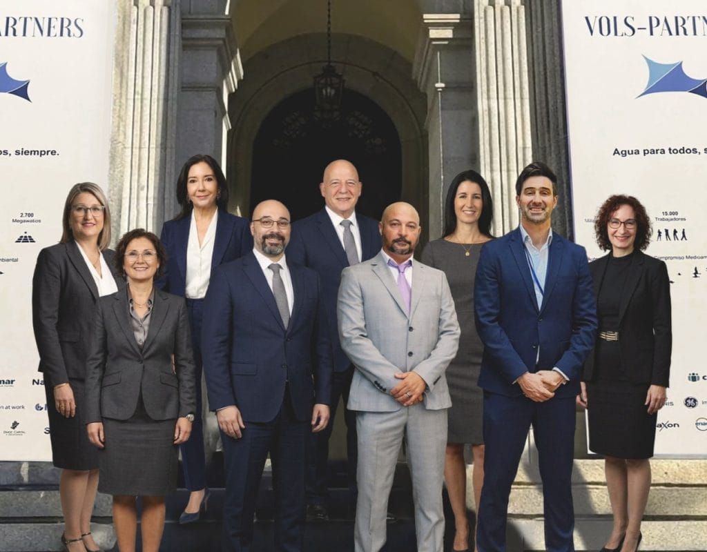 Eight smiling professionals in formal business attire pose on steps in front of an arched doorway, flanked by white banners.&nbsp;Hispamer Noticias