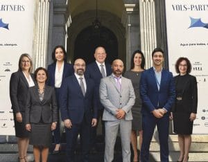 Eight smiling professionals in formal business attire pose on steps in front of an arched doorway, flanked by white banners.&nbsp;Hispamer Noticias