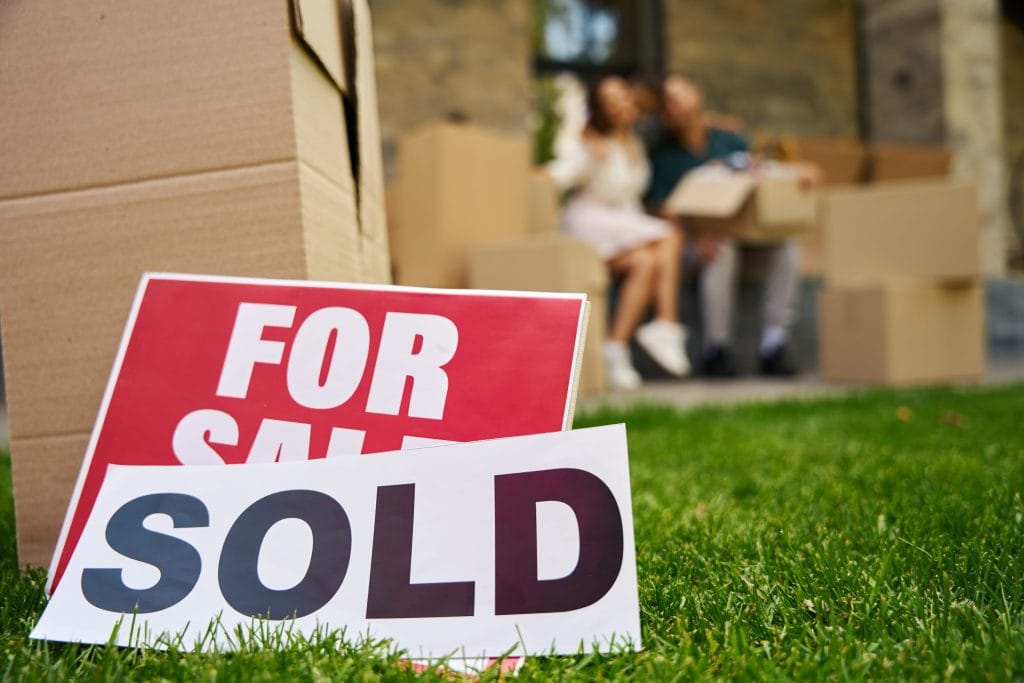 Real estate signs in the foreground read 'FOR SALE' with a 'SOLD' sign overlapping, on a grassy yard with moving boxes and a couple in the background.&nbsp;Hispamer Noticias