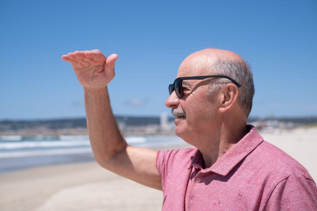 Older man in a pink polo shirt and sunglasses stands on a sunny beach, shielding his eyes with his hand.&nbsp;Hispamer Noticias