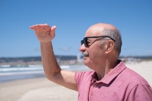 Older man in a pink polo shirt and sunglasses stands on a sunny beach, shielding his eyes with his hand.&nbsp;Hispamer Noticias