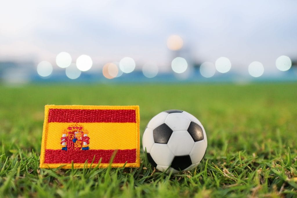 Spanish flag patch beside a soccer ball on green grass with blurred stadium lights in the background&nbsp;Hispamer Noticias