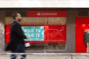 Santander bank storefront with a blurred pedestrian in the foreground; a turquoise billboard in the window reads 'PORQUE TÚ'. When in context, conveys a bank branch with a bold blue ad.&nbsp;Hispamer Noticias