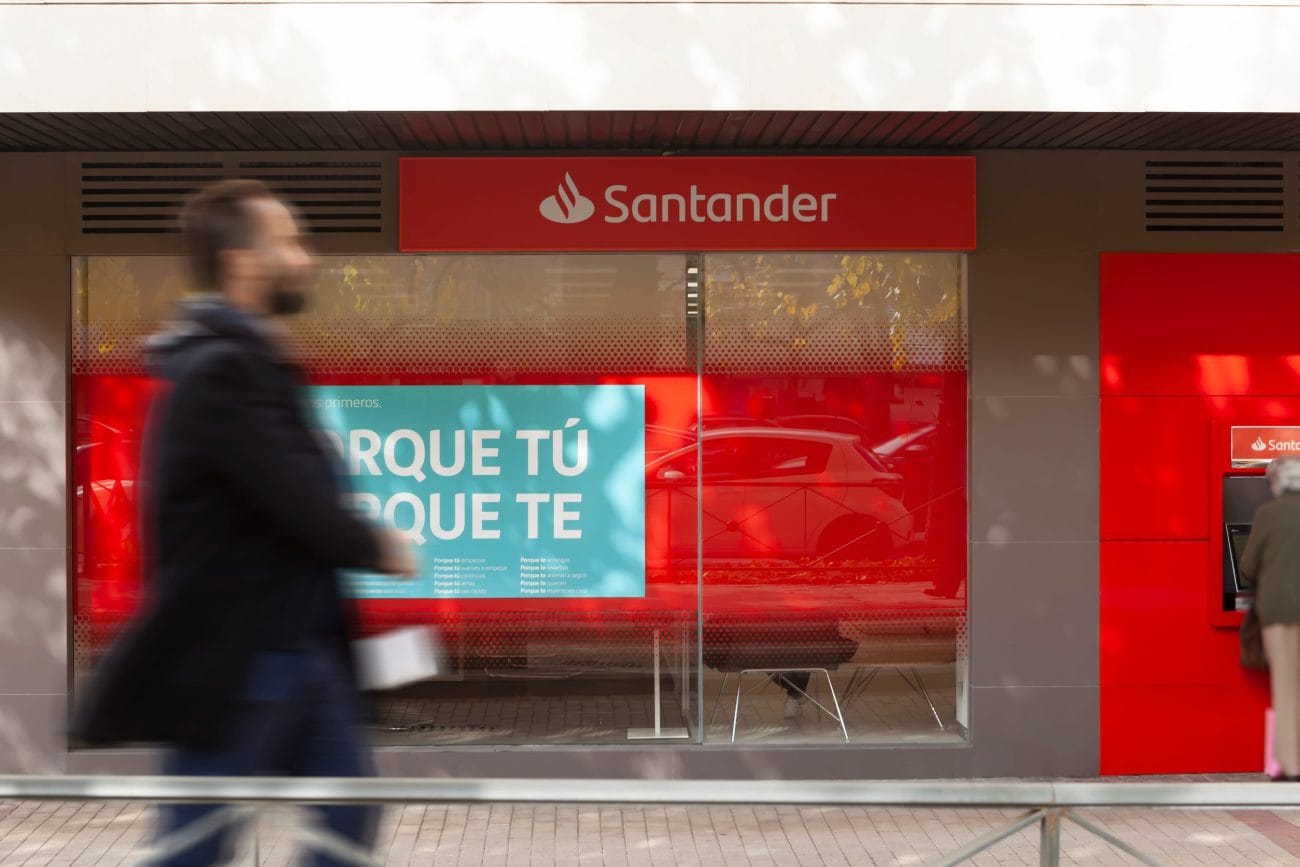 Santander bank storefront with a blurred pedestrian in the foreground; a turquoise billboard in the window reads 'PORQUE TÚ'. When in context, conveys a bank branch with a bold blue ad.&nbsp;Hispamer Noticias