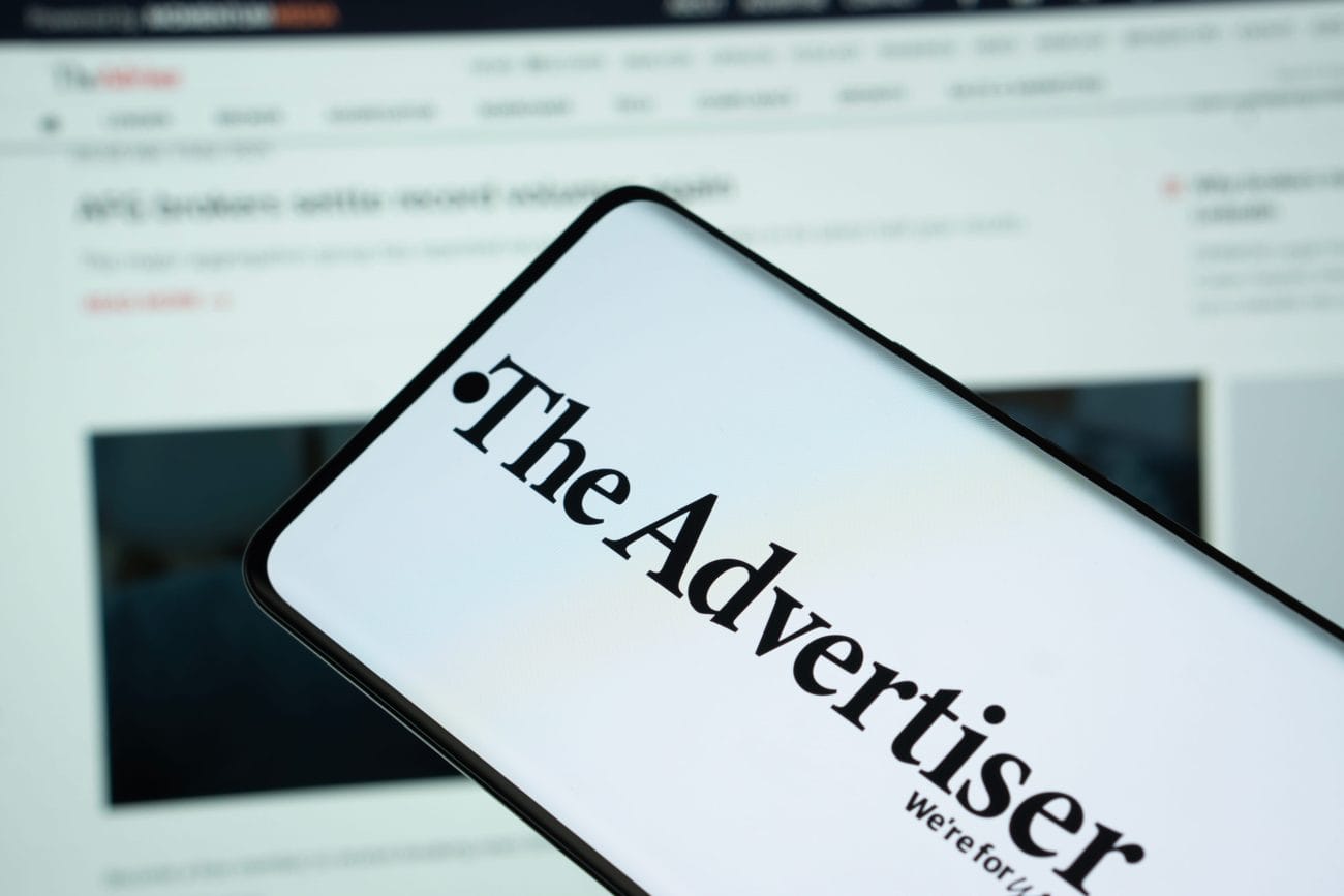 campaña publicidad - Hispamer Noticias Close-up of a smartphone showing the bold 'The Advertiser' logo, held over a blurred computer screen displaying a website in the background. Hispamer Noticias