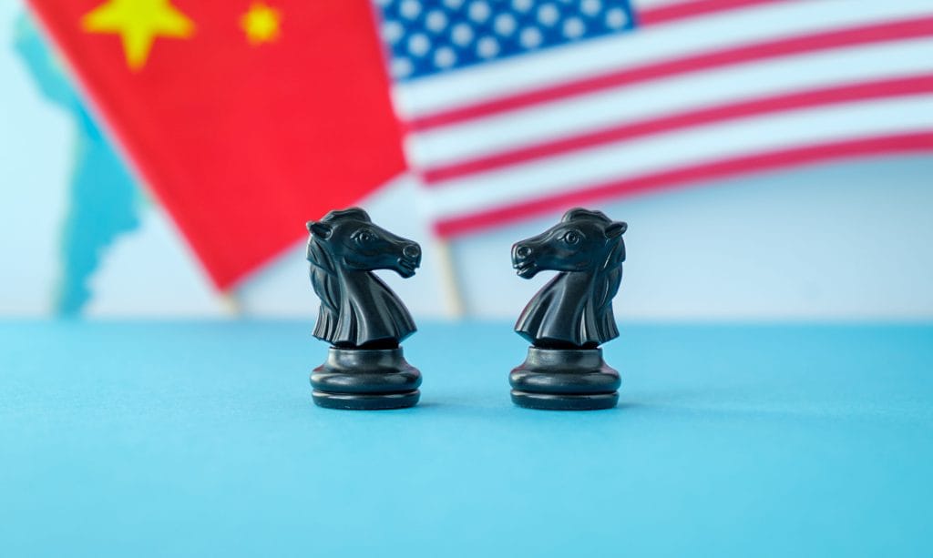 Two black knight chess pieces facing each other on a blue surface with blurred flags of China and the United States in the background, suggesting competition or diplomacy.&nbsp;Hispamer Noticias