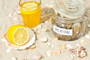 Jar labeled VACATION filled with coins sits on beach sand beside a glass of lemonade and a halved lemon on a shell, with seashells around.&nbsp;Hispamer Noticias