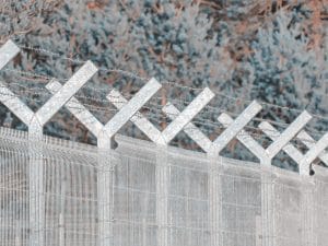 Metal security fence with barbed wire along the top and V-shaped support posts, outdoors against shrubs and trees behind.&nbsp;Hispamer Noticias