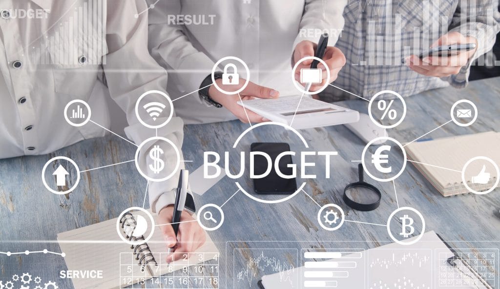 Illustration of budgeting concept with interconnected icons (currency, charts, lock, percentage) and the word BUDGET at center on a desk scene.&nbsp;Hispamer Noticias