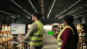 Two warehouse workers in high-visibility vests monitor inventory on a desk computer while a colleague checks a tablet in a storage facility?docks?&nbsp;Hispamer Noticias