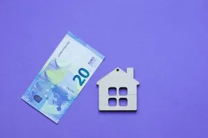 Twenty-euro banknote next to a wooden house icon on a purple background, illustrating housing costs or mortgage concepts.&nbsp;Hispamer Noticias