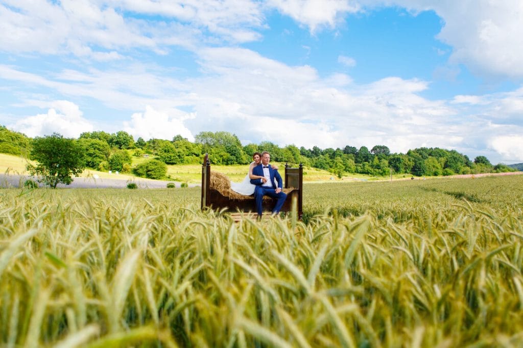 Bride and groom sit on a wooden bed in a golden wheat field beneath a bright blue sky, posing for a wedding photo.&nbsp;Hispamer Noticias