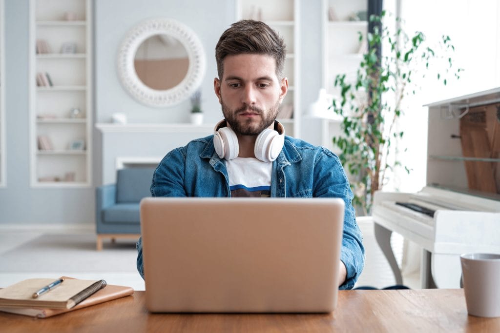 Bearded man wearing white over-ear headphones works on a laptop at a wooden table in a bright living room.&nbsp;Hispamer Noticias