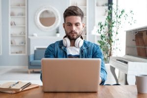 Handsome modern man designer working on laptop online using internet at home - Hispamer Noticias Bearded man wearing white over-ear headphones works on a laptop at a wooden table in a bright living room. Hispamer Noticias