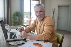 Older man with gray hair and beard sits at a wooden table with an open laptop, holding glasses and smiling at the camera amid papers and charts nearby, in a home setting.&nbsp;Hispamer Noticias
