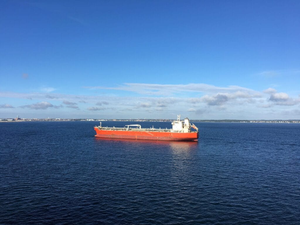 A large red cargo ship sails on calm blue water with a distant shoreline on the horizon under a clear sky.&nbsp;Hispamer Noticias