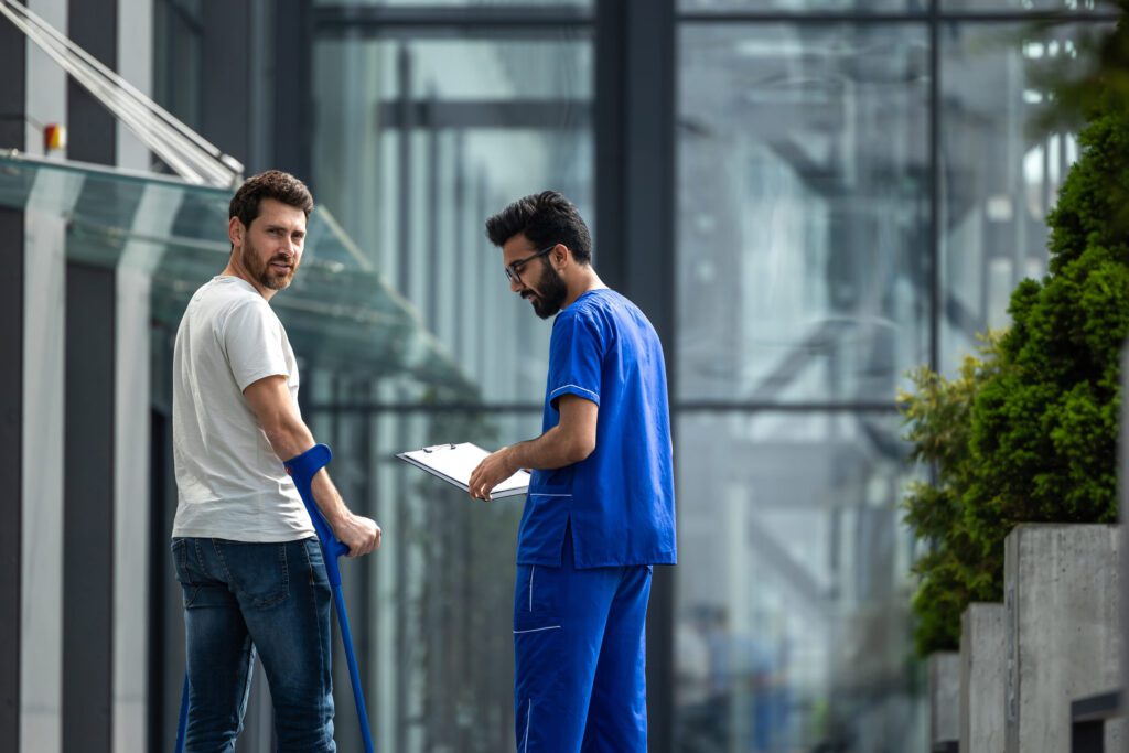 Man with a blue forearm crutch speaks with a clinician in blue scrubs who holds a clipboard outside a glass-walled building.&nbsp;Hispamer Noticias